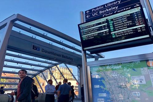 Downtown Berkeley station entrance with signage including a transit route map, live transit updates on digital screen, and signange pointing to various points of interest. 