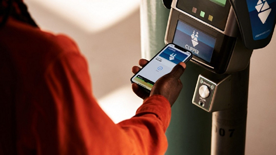 Person holding up mobile device with Clipper application open, to a Clipper kiosk to pay ride fare. 