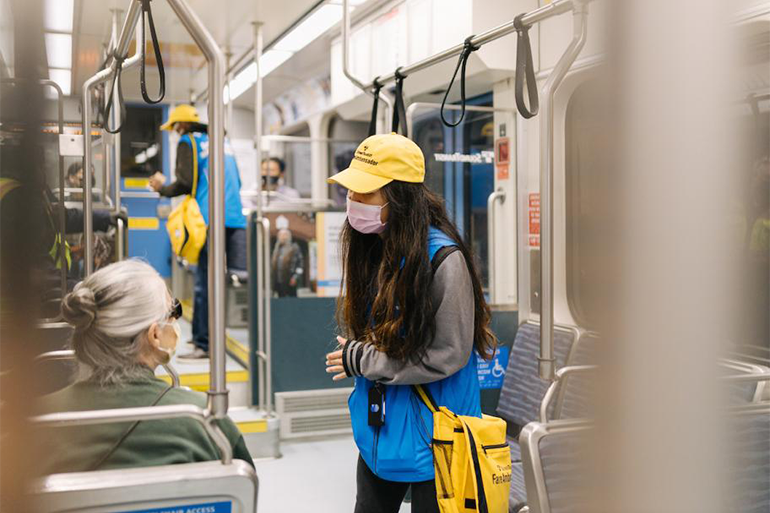 Two fare ambassadors in new uniforms, including yellow hats and bags and blue vests, assisting passengers on a Link light rail train.