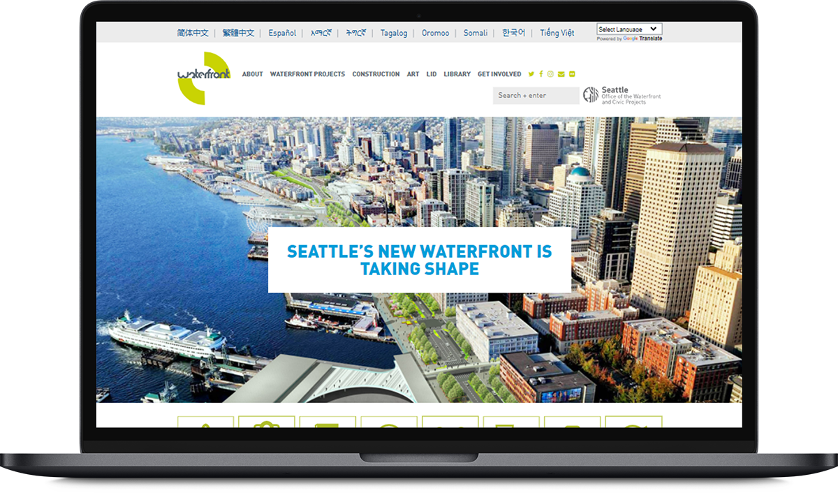 A laptop is opened with the screen showing an aerial image of the Seattle Waterfront participate-online website. The banner on the home Page is shown. On the left side of the banner image is the blue water of the Puget Sound; a ferry can be seen docking at the terminal. To the right is downtown Seattle, with tall skyscrapers. In the center of the image the downtown core is separated by smaller mixed-use buildings, greenery and trees. Overlaid in the center of the banner is the text that reads “Seattle’s New Waterfront is Taking Shape.” 