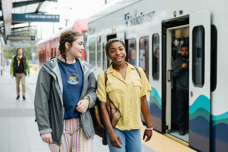 Two passengers--one with a gray sweater and a blue shirt and one with a yellow shirt and brown backpack--walking alongside a Link light rail train.