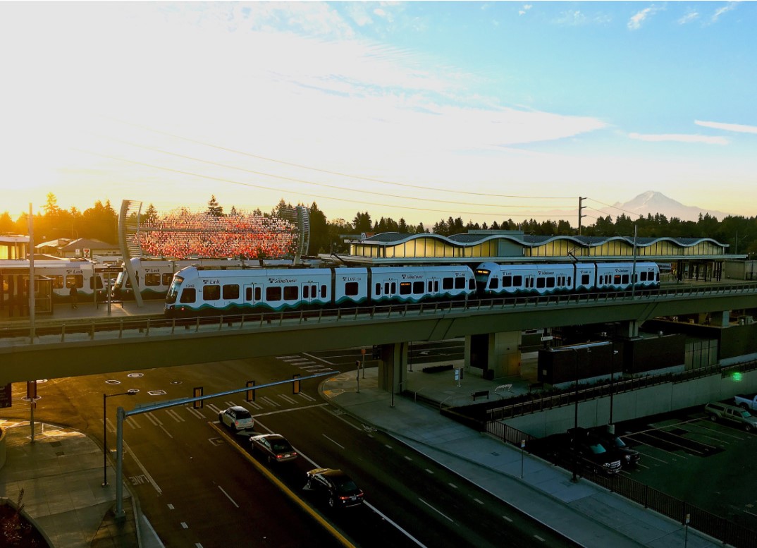 Mount Rainier를 배경으로 이른 아침 해뜰 무렵 기존 경전철 역에 도착하는 Link 경전철 열차를 보여주는 사진. 역은 지상에서 올라가고 철도는 지상에서 운행됩니다. 사진은 SR 526 Evergreen에 건설될 것과 같은 고가 역이 어떤 모습일지 예시를 보여줍니다.