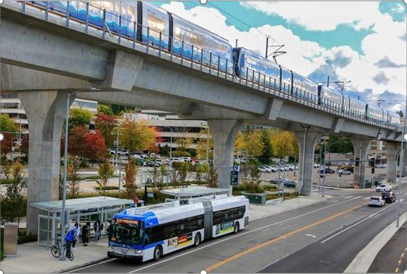 파란색과 흰색의 Link 경전철 열차는 파란색의 흐린 하늘을 배경으로 강조 표시된 높은 콘크리트 트랙을 따라 이동합니다. 파란색과 흰색의 Sound Transit 버스 아래 양방향 도로에서 5명의 보행자가 탑승을 기다리는 버스 정류장에 있습니다. 배경에서 사무실 건물과 주차장은 다양한 녹색, 노란색 및 빨간색 나무를 통해 볼 수 있습니다. 

                          사진은 Everett에 건설될 고가 역이 어떤 모습일지 보여줍니다.
